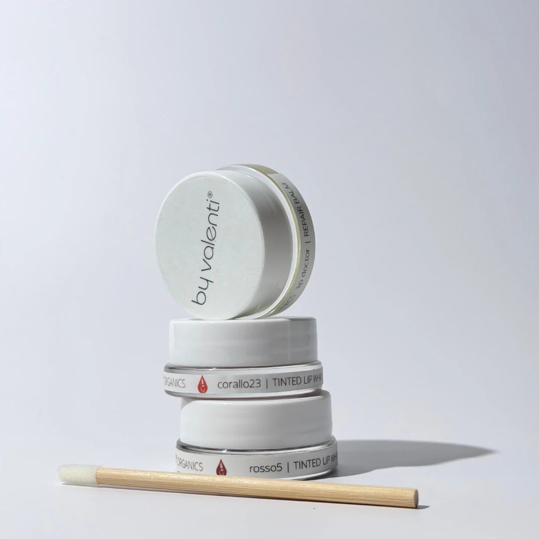Stack of By Valenti Tinted Whipped Lip Balms containers and a lip wand with 'by valenti' branding on a light gray background