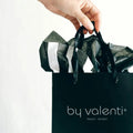 Hand holding a black 'by valenti' shopping bag with ribbons against a white background