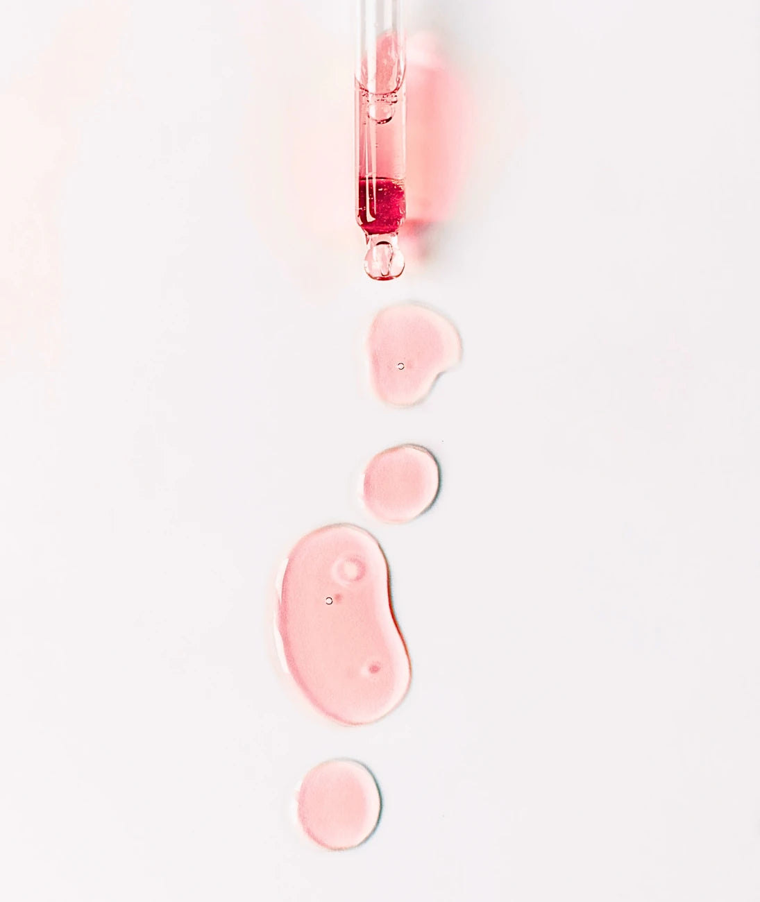 By Valenti pink skincare serum spilling on a white surface from a glass dropper tip